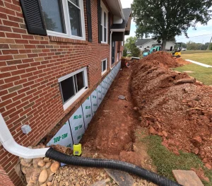 Brick house exterior with a trench along the foundation, exposing waterproofing, soil piles, and scattered construction tools. A calm, work-in-progress vibe.