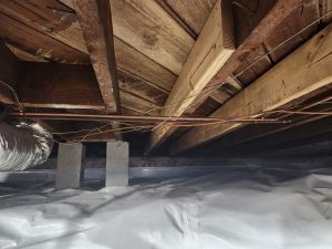 Wooden ceiling beams in a crawl space with visible wiring and a metal duct. Concrete blocks and a white plastic ground cover add an unfinished look.